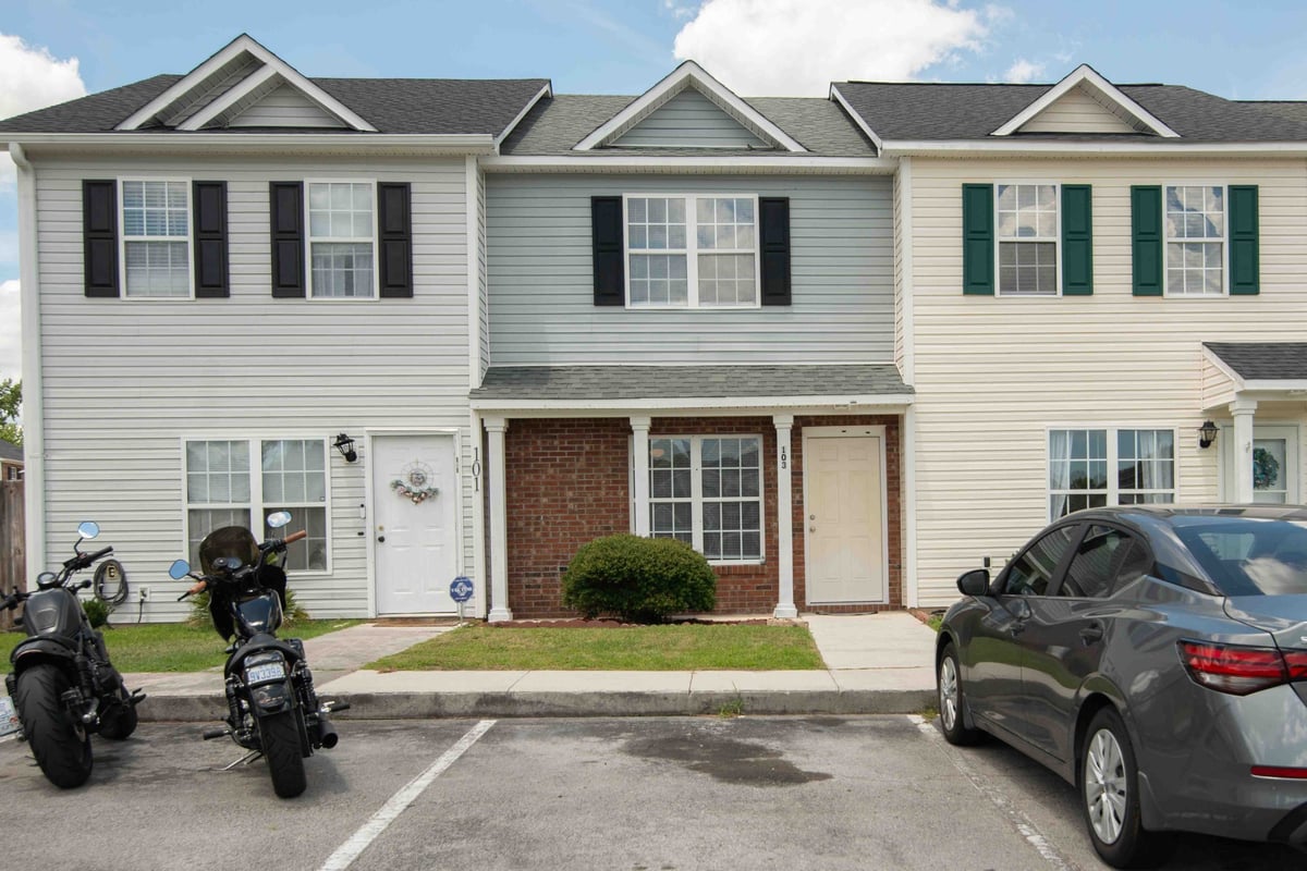 Row of townhouses with white and gray vinyl siding, black shutters, brick front entry, parked motorcycles and sedan in driveway on sunny day